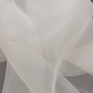 Puede incluir: Una tela blanca transparente y ligera con una textura fina y traslúcida. El material está drapeado en pliegues suaves y fluidos, creando variaciones sutiles en la opacidad y el reflejo de la luz sobre la superficie lisa y delicada.