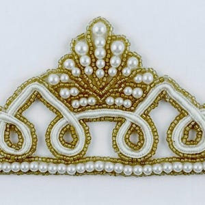 Puede incluir: Una aplicación en forma de corona con una base de cordón blanco, cuentas doradas y perlas artificiales blancas. El diseño incluye un pico central elevado con perlas y una fila horizontal de perlas a lo largo del borde inferior.