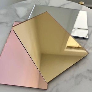 Può includere: Tre lastre acriliche riflettenti in oro, argento e oro rosa. Le lastre sono rettangolari e disposte ad angolazioni diverse, riflettendo la luce e creando un'estetica geometrica moderna. Le superfici sono lisce e lucide.
