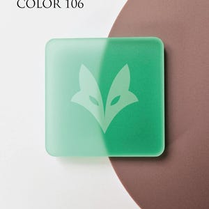 Op de afbeelding: Een vierkant, tweekleurig, mintgroen object met afgeronde hoeken. De linkerkant is een lichtere tint groen dan de rechterkant. Een wit vossenkoplogo is gecentreerd op het object. De tekst "COLOR 106" staat in de linkerbovenhoek.