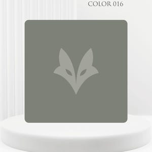 Può includere: Un oggetto quadrato verde oliva con un logo stilizzato a forma di testa di volpe al centro. Il testo "COLOR 016" è visibile. L'oggetto è presentato su un piedistallo bianco arrotondato su sfondo bianco.