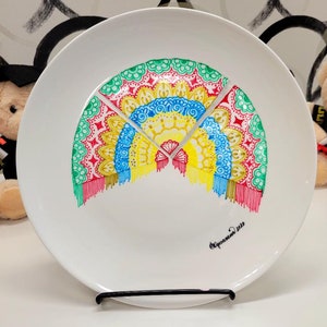 Könnte beinhalten: Ein weißer Keramikteller mit einem handgemalten Design eines bunten Fächers mit roten, gelben, blauen, grünen und weißen Details. Der Teller hat einen schwarzen Ständer und der Text "Oprahwind 2022" ist unten aufgedruckt.