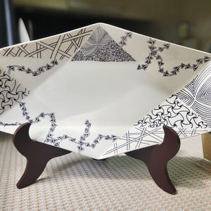 Könnte beinhalten: Eine weiße Keramik-Servierplatte mit sechseckiger Form und schwarzen Tinten-Kritzel-Designs. Die Platte steht auf einem dunkelbraunen Holzständer.