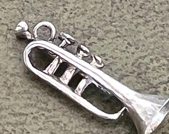 May include: A silver-toned metal trumpet charm featuring a detailed mouthpiece, three valves, and a flared bell. The musical instrument pendant includes a small circular jump ring at the top for attachment to a chain or bracelet, with a polished finish.
