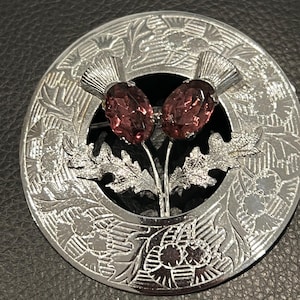 May include: A circular silver-tone brooch featuring an embossed thistle pattern on the outer rim. The center displays two metallic thistle stems topped with oval-shaped, faceted purple glass stones and detailed silver-tone leaves at the base.