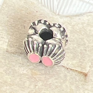 May include: A silver charm with a heart-shaped design and a black center. The charm features a ribbed texture and two pink enamel circles at the base. The charm is likely for a bracelet or necklace.