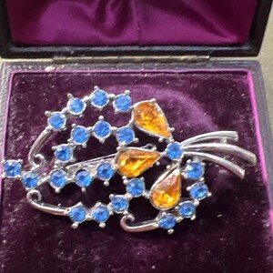 Puede incluir: Broche vintage plateado con diseño floral. El broche presenta piedras preciosas azules y ámbar dispuestas en un arreglo estilizado. El broche se exhibe en una caja forrada de terciopelo morado.
