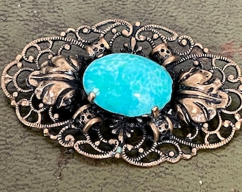 May include: An oval-shaped brooch featuring a central, smooth, bright turquoise cabochon stone. The stone is surrounded by an intricate, dark-toned metal filigree frame with detailed leaf-like motifs on the left and right sides of the setting.