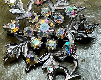Könnte beinhalten: Silberfarbene Brosche in floralem Design mit irisierenden, runden und ovalen Strasssteinen. Die Brosche hat ein detailliertes Blatt- und Stielmuster, wobei die Strasssteine in Größe und Farbe variieren und einen funkelnden Effekt erzeugen.