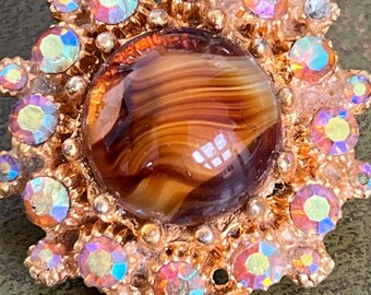 May include: A vintage gold-toned brooch with a central, marbled brown stone. The brooch is surrounded by iridescent, colorful rhinestones and smaller gold accents, creating a sunburst design. The brooch is a classic piece of jewelry.