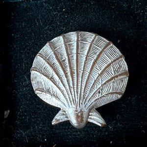 May include: A silver-toned metal brooch shaped like a scallop shell. The piece features a textured, fan-like design with radiating ridges and grooves that extend from the base to the rounded top edge, creating a detailed, realistic shell appearance.
