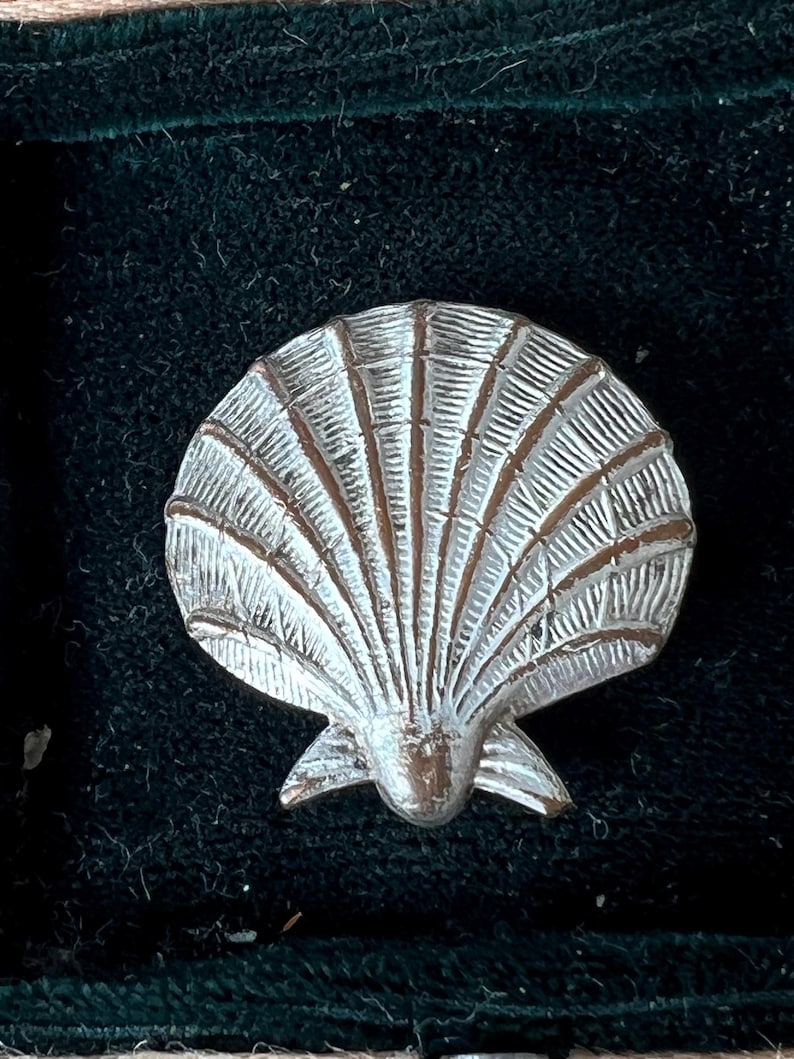 May include: A silver-toned metal brooch shaped like a scallop shell. The piece features a textured, fan-like design with radiating ridges and grooves that extend from the base to the rounded top edge, creating a detailed, realistic shell appearance.