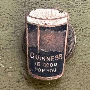 May include: A vintage Guinness pin featuring a pint of stout with a white foamy head. The pin has a dark blue section with the text "Guinness is good for you". The pin is set on a silver-colored metal base.