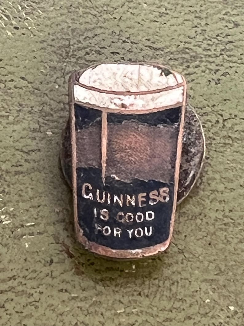 May include: A vintage Guinness pin featuring a pint of stout with a white foamy head. The pin has a dark blue section with the text "Guinness is good for you". The pin is set on a silver-colored metal base.