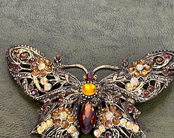 Könnte beinhalten: Eine schmetterlingsförmige Brosche aus dunklem, antikisiertem Metall mit filigranen Mustern. Die Flügel sind mit einer Ansammlung von bernsteinfarbenen, braunen und klaren facettierten Kristallen verziert, mit einem runden Bernstein in der Mitte.