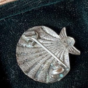 May include: A silver-toned metal brooch shaped like a scallop shell with a textured, radiating ribbed surface. The back features a simple pin mechanism with a hinged clasp and a catch, showing signs of oxidation and wear on the metallic finish.