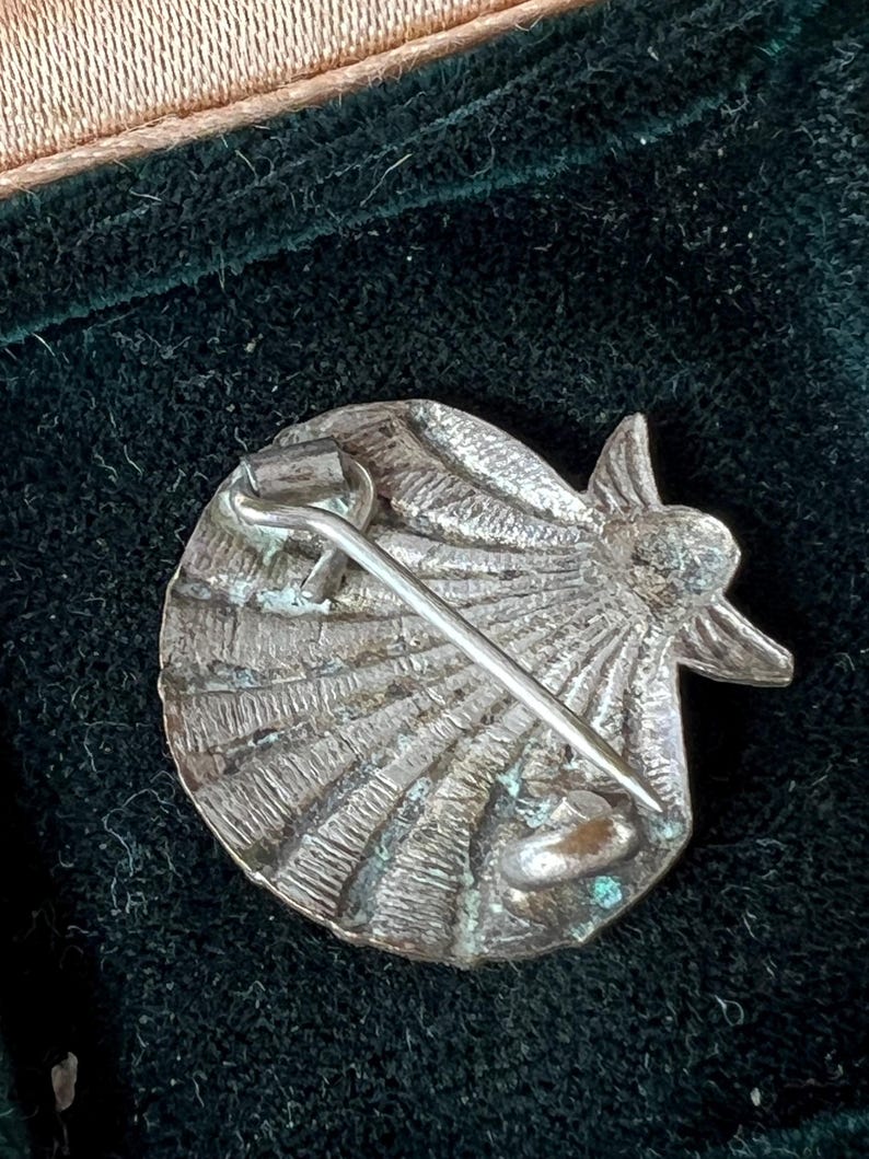 May include: A silver-toned metal brooch shaped like a scallop shell with a textured, radiating ribbed surface. The back features a simple pin mechanism with a hinged clasp and a catch, showing signs of oxidation and wear on the metallic finish.
