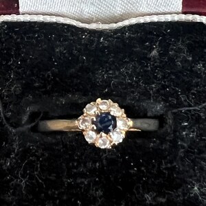 May include: A gold ring featuring a central round dark blue gemstone surrounded by a cluster of ten small clear white stones in a floral arrangement. The ring is set against a black velvet interior within a dark red jewelry box.