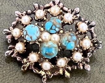 Könnte beinhalten: Eine kreisförmige Brosche mit einem texturierten, dunkelmetallischen Rahmen, verziert mit vier runden, blau geäderten Türkis-Cabochons in Kreuzform, durchsetzt mit kleinen, runden, weißen Perlen, die in das kunstvolle Metallwerk eingefasst sind.