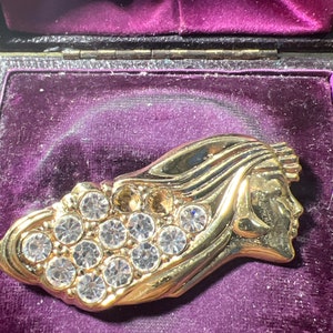 May include: A gold-toned brooch featuring a stylised female profile with a flowing hair design. The brooch is adorned with clear rhinestones.
