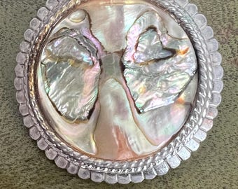 May include: A circular brooch featuring a central inlay of iridescent abalone shell with pink, green, and cream tones. The shell is surrounded by a twisted rope-style metal border and an outer scalloped edge, all finished in a polished silver tone.