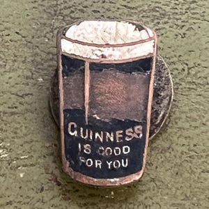 May include: A vintage pin featuring a pint of Guinness stout. The pin has a white foamy top, a dark brown body, and a black label with the text "Guinness is good for you" in white lettering. The pin is set against a green background.