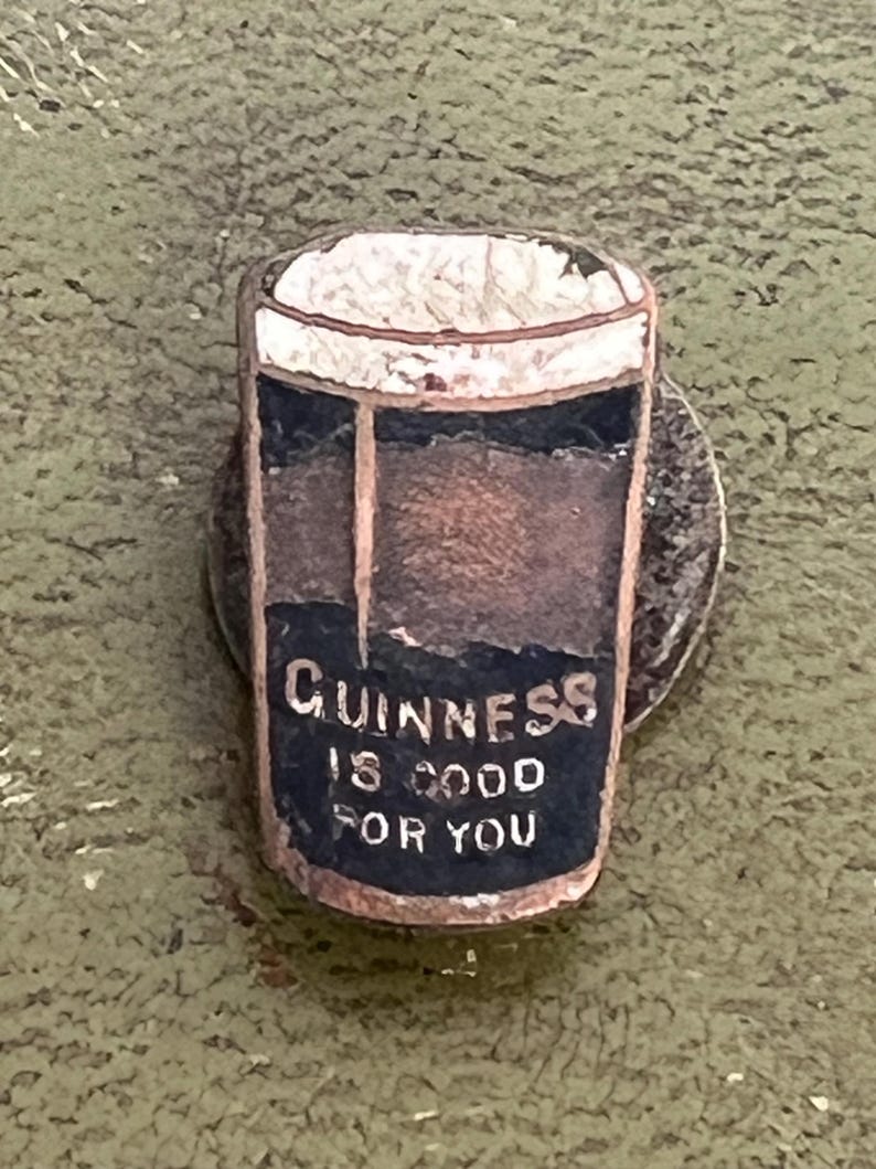 May include: A vintage pin featuring a pint of Guinness stout. The pin has a white foamy top, a dark brown body, and a black label with the text "Guinness is good for you" in white lettering. The pin is set against a green background.