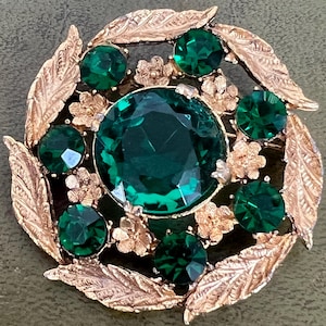 May include: A round brooch featuring a large central emerald green faceted stone surrounded by six smaller matching green stones. The frame is crafted from gold-toned metal with a textured leaf pattern and small floral accents between the stones.