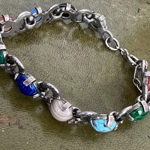 May include: A silver-toned link bracelet featuring a series of alternating polished stones in various colours including blue, green, red, and banded white. The metal links are textured and connected by small loops, with a secure clasp mechanism visible.