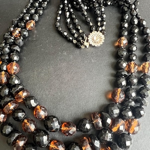 Può includere: Una collana a più fili con perline sfaccettate nere e color ambra traslucido. La collana è composta da diversi strati di perline e include una chiusura rotonda in metallo color argento con un design floreale sul gancio.