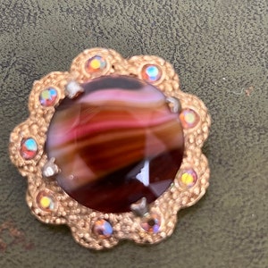 Op de afbeelding: Een gouden broche met een grote, ronde, bruine en roze agaat-steen, omringd door kleinere, iriserende stenen.