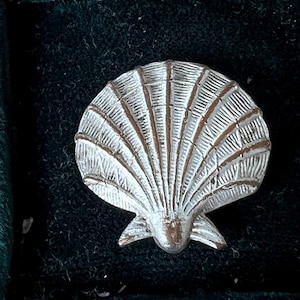 May include: A silver-toned metal brooch shaped like a scallop shell. The surface features raised, radiating ridges with a textured, weathered finish in the recessed areas, creating a distinct contrast against the polished metallic highlights of the shell.