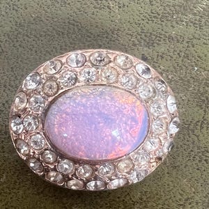 Könnte beinhalten: Eine runde, silberfarbene Brosche mit einem großen, schillernden ovalen Stein, der von einem Heiligenschein aus klaren Strasssteinen umgeben ist.