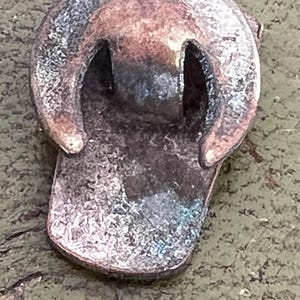 May include: Close-up of a weathered, metallic object with a rounded top and a rectangular base. The object has two arched openings and a distressed, aged appearance with shades of brown, copper, and hints of blue-green. The texture appears rough and worn.
