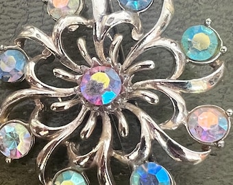 May include: A silver-toned brooch with a floral design. The brooch features a central iridescent stone surrounded by eight smaller iridescent stones. The design has curved, leaf-like elements. The stones reflect a range of colors, including blue, pink, and yellow.