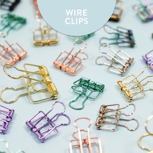 Pode incluir: Uma variedade de clipes de encadernação de arame coloridos, incluindo ouro, prata, ouro rosa, verde, roxo e iridescente. Os clipes estão dispostos sobre um fundo azul claro. O texto "WIRE CLIPS" está escrito em branco sobre um círculo azul claro.