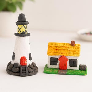 May include: Miniature lighthouse and house figurines. The lighthouse is white with black accents, a red door, and a black top. The house has a yellow roof, a red door, and sits on a green base. These figurines are ideal for a coastal or nautical theme.