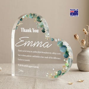 May include: A heart-shaped clear acrylic plaque featuring embedded green eucalyptus leaves and gold flakes along the curved edge. The text reads: Thank You Emma, followed by a message of gratitude and the name John, printed in white lettering.