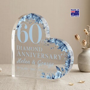 May include: A clear acrylic heart-shaped anniversary keepsake with blue floral accents. The text reads "60th Diamond Anniversary Helen & George" in light blue. A small vase with dried flowers is in the background. Made in Australia.