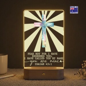 May include: Illuminated cross-shaped night light with a watercolour design. The light features a cross in shades of pink and blue against a background of radiating lines. The text reads "FEAR NOT FOR I HAVE REDEEMED YOU; I HAVE CALLED YOU BY NAME you are mine ISAIAH 43:1".