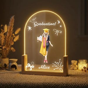 May include: An illuminated graduation-themed night light. It features a graduate figure, a cap, and gown, holding a champagne glass. The words "Graduation!" and "Alice" are displayed. The light has a wooden base and a warm, inviting glow.