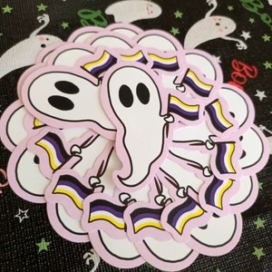 May include: A stack of stickers featuring a cartoon ghost holding a pride flag with yellow, purple and black stripes. The ghost is pink with black eyes and a pink outline. The stickers are on a black background with white stars.