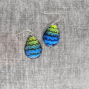 May include: A pair of teardrop-shaped earrings with a blue, green, and black abstract pattern. The earrings have silver hooks.