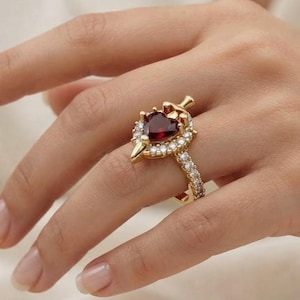 May include: A gold ring featuring a central heart-shaped dark red gemstone surrounded by a halo of small clear stones. The band is also encrusted with small clear stones, and the setting includes a small gold dagger-like accent at the top.