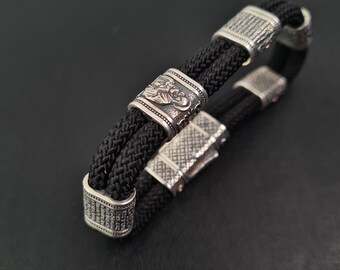 May include: Black braided bracelet with silver metal detailing. The bracelet has a woven design with decorative metal pieces and a clasp. The metal pieces feature intricate patterns and textures, adding detail to the accessory.