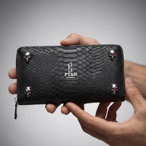 May include: A black rectangular wallet with a textured snakeskin pattern. The front features four silver-toned skull ornaments with red eyes at the corners and the silver text PTAH jewelry centered. A metal zipper runs along the top edge.