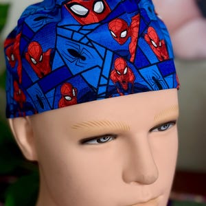 Op de afbeelding: Een blauwe chirurgische scrub cap met een herhalend patroon van Spider-Man. Het ontwerp bevat Spider-Mans gezicht en volledige lichaam in rood, met een blauwe achtergrond en zwarte spinnenweb accenten.