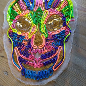 Peut inclure: Un écusson de crâne coloré avec des broderies vert néon, rose, bleu et orange. Les yeux et le nez du crâne sont remplis de paillettes dorées. L'écusson est sur un tissu transparent blanc cassé.