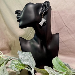 Może przedstawiać: Srebrne i czarne kolczyki w kształcie kawiarki ze złotymi haczykami. Kolczyki mają kształt klasycznego ekspresu do kawy. Kolczyki są prezentowane na czarnej głowie manekina na neutralnym tle.