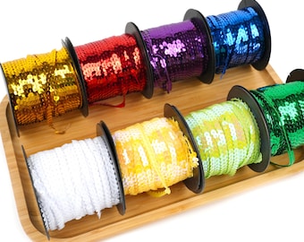 May include: A set of eight spools of colorful sequined trim, arranged on a wooden tray. The trim is in various colors, including gold, red, purple, blue, green, and white.